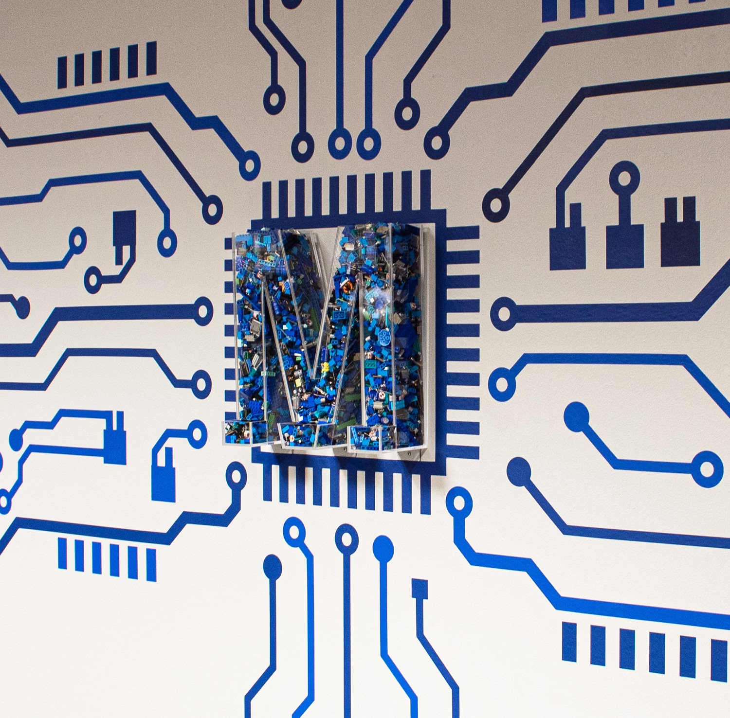 Decals of circuit lines all pointing to an acrylic 3-D letter M that is filled with blue legos and electronic components